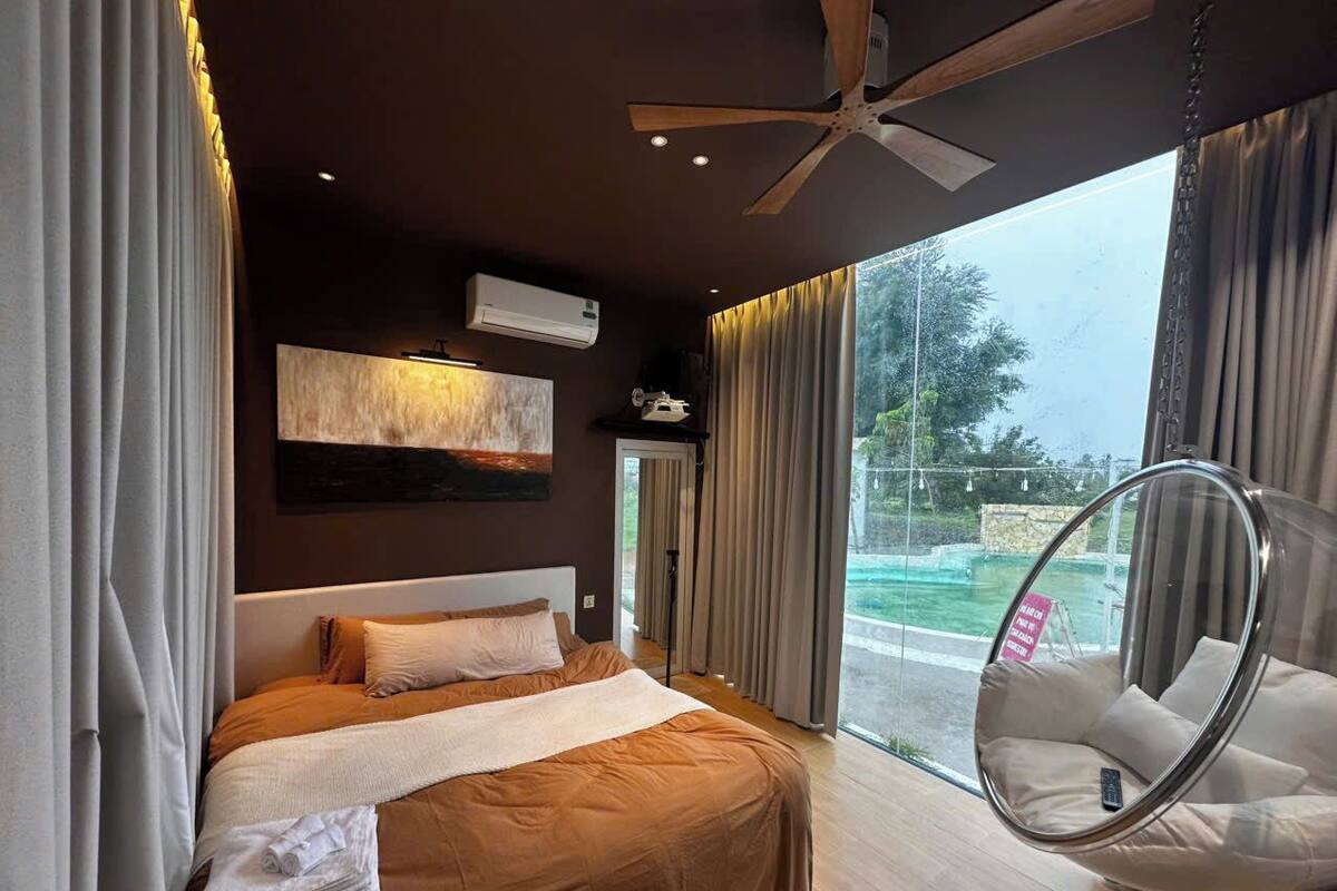 A bedroom with a bed, a hanging chair, a ceiling fan, and a view of the outdoors.