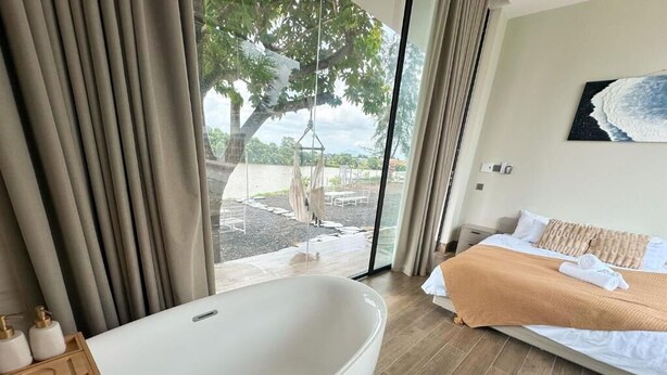 Un dormitorio con una bañera grande, una cama con una manta color beige, y vistas a un árbol y al agua desde el exterior.