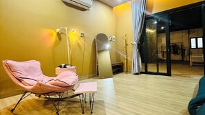 Una habitación con una silla rosa, una pequeña mesa, un equipo de aire acondicionado y una puerta de cristal que da al exterior.