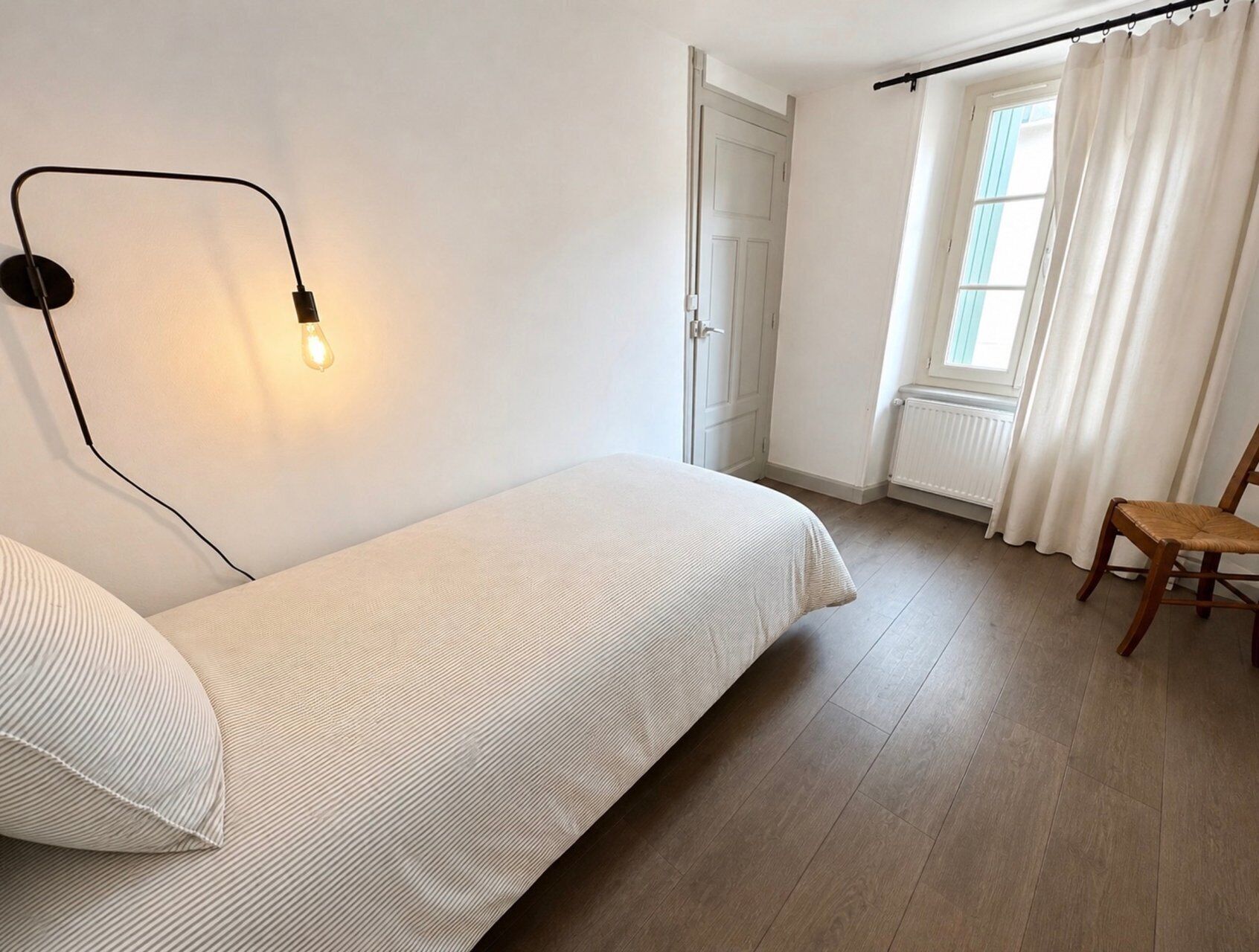 Et soveværelse med seng, stol, et vindue med gardiner og en vægmonteret lampe.