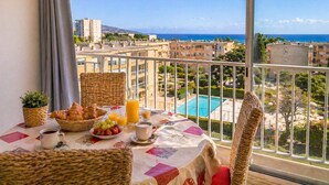 Een balkon met ontbijt, onder meer croissants, fruit en sap, met uitzicht op een woongebied en de zee.