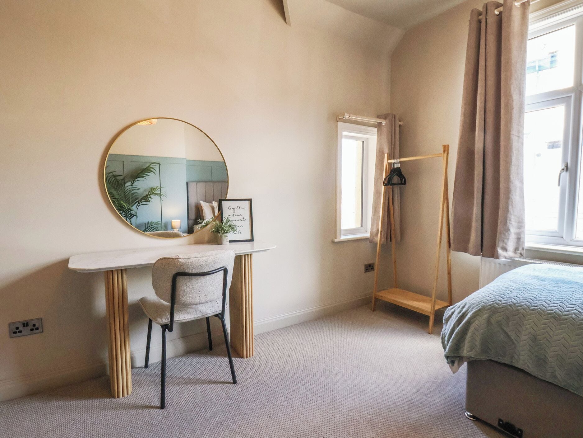 A bedroom with a round mirror, a desk, a chair, a bed, a window with curtains, and a radiator.