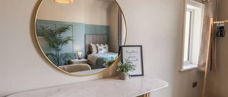 A bedroom with a large mirror, a bedside table with a lamp, a framed inspirational quote, and a window with curtains.