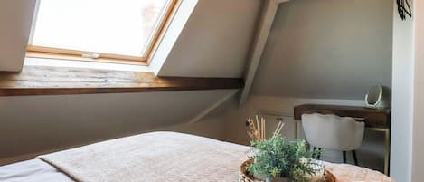 A bedroom with a skylight, a bed with a textured blanket, a small desk, and a potted plant.