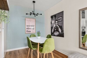 Een eetruimte met een ronde tafel, groene stoelen, een hanglamp, een ingelijste citaat aan de muur en een groot, ingelijst schilderij.