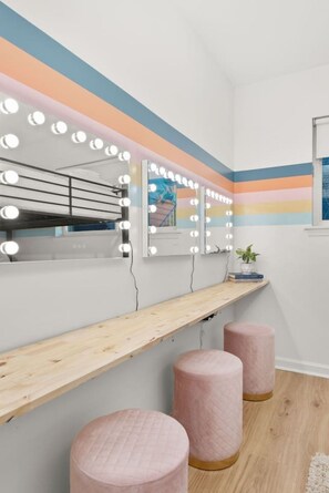 Ein Zimmer mit Schminkbereich, ausgestattet mit einem Spiegel mit Beleuchtung, einer Holzablage und zwei rosa Hocker.