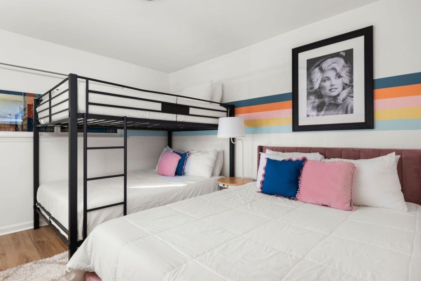 Una habitación con literas, una mesita de noche con lámpara y una pared de rayas coloridas.