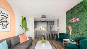 Ein modernes Wohnzimmer mit Sofa, Couchtisch und Sesseln, eine Küche mit Barhocker und eine grüne Wand mit einem Neon-Leuchtschild.