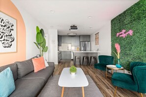 A modern living room with a sofa, coffee table, and chairs, a kitchen with bar stools, and a green wall with a neon sign. - Best Location! Walk to Midtown Bars (Nashville)