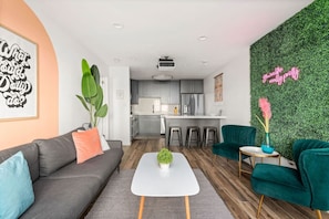 Ein modernes Wohnzimmer mit Sofa, Couchtisch und Sesseln, eine Küche mit Barhocker und eine grüne Wand mit einem Neon-Leuchtschild.