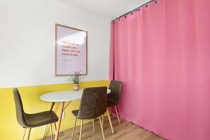 Una habitación con una mesa blanca, dos sillas, una cortina rosa y una cita enmarcada en la pared.