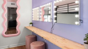 Ein Zimmer mit Schminktisch, zwei rosafarbenen Hocker, einem Holzregal und einem gewellten Spiegel mit Beleuchtung.