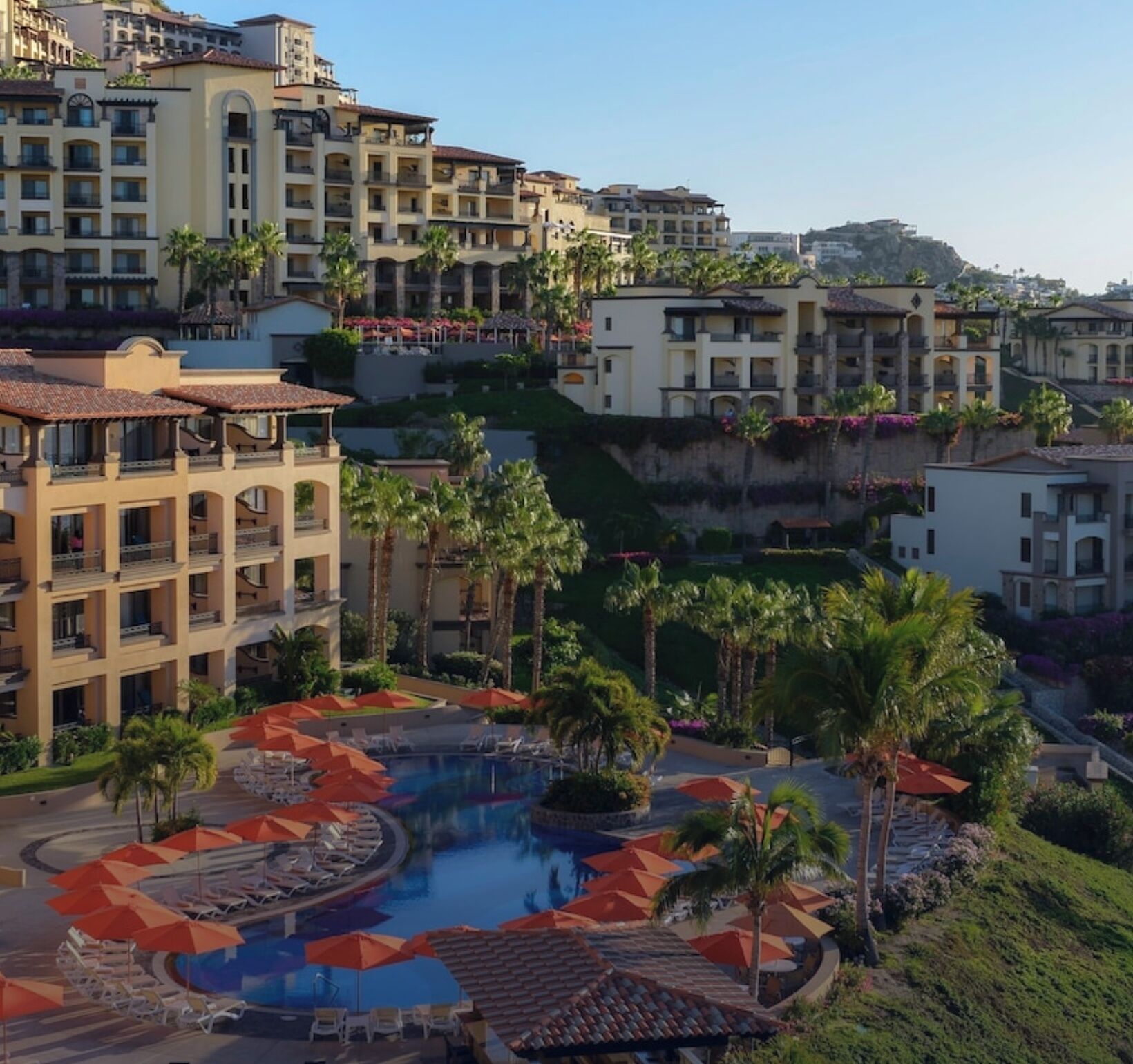 Un resort con piscina central rodeada de tumbonas y sombrillas, varias construcciones con balcones y exuberante vegetación.
