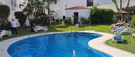A swimming pool with lounge chairs, a covered area, and a building in the background.