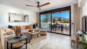 Una sala moderna con sofá, mesa de centro y reposapiés, ventilador de techo y un amplio ventanal corredera que da a un área de comedor exterior con vista a la montaña.