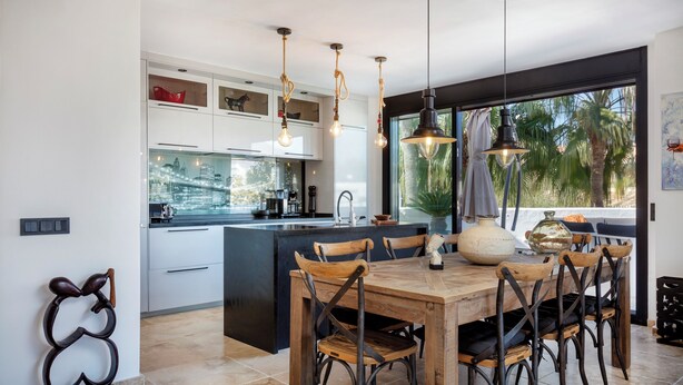 Una cocina moderna con una isla central, luces colgantes y un comedor con sillas de madera.