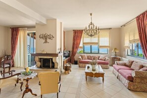 A living room with a fireplace, sofas, a chandelier, and a view of the sea. - Holiday Home 'Villa Oneiro' with Sea View, Wi-Fi and Air Conditioning (Στούπα)