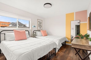 Une chambre avec deux lits, un bureau, un miroir et une fenêtre donnant sur des immeubles.