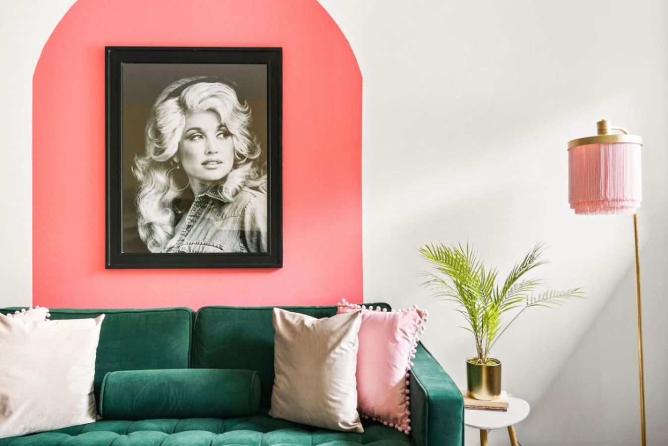 Ein grünes Sofa mit einem gerahmten Bild darüber, ein kleiner runder Tisch mit einer Pflanze und eine hohe Stehlampe mit einem rosa Lampenschirm.