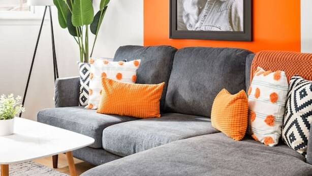 A grey sectional sofa with orange and patterned cushions, a framed portrait on an orange wall, and a large green plant. - The Lyric - Music Row Right at Your Doorstep (Nashville)