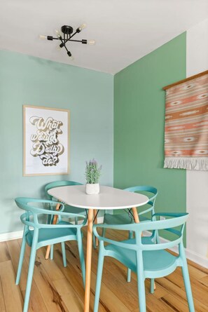 Un coin repas avec des chaises de couleur turquoise, une table blanche, un cadre contenant une citation, une plante en pot et un rideau à motifs.