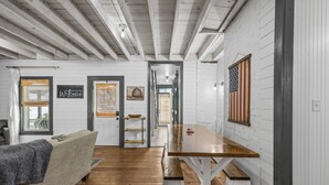 Une pièce avec un plancher en bois, un canapé blanc, une table à manger en bois et un banc. Il y a deux portes, l’une donnant sur une cuisine et l’autre sur un couloir.