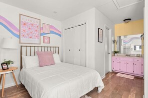 Ein Schlafzimmer mit einem Bett, einem Nachttisch mit Lampe, einem rosa Kissen, einem gerahmten Bild und einem rosa Schminktisch mit Spiegel.