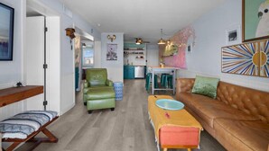 Ein Wohnzimmer mit einer Couch, einem Sessel, einem Tisch und einer Bank. Im Hintergrund ist eine Küche zu sehen.