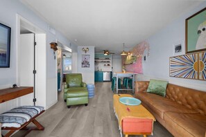 Ein Wohnzimmer mit einer Couch, einem Sessel, einem Tisch und einer Bank. Im Hintergrund ist eine Küche zu sehen.