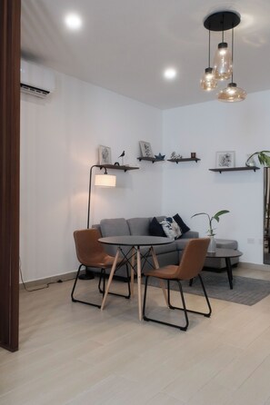Ein modernes Wohnzimmer mit einem grauen Sofa, zwei braunen Sesseln, einem kleinen runden Tisch und einer Pendelleuchte. An der Wand befinden sich Regale mit Dekoration, und ein Stehschrank sorgt für Licht.