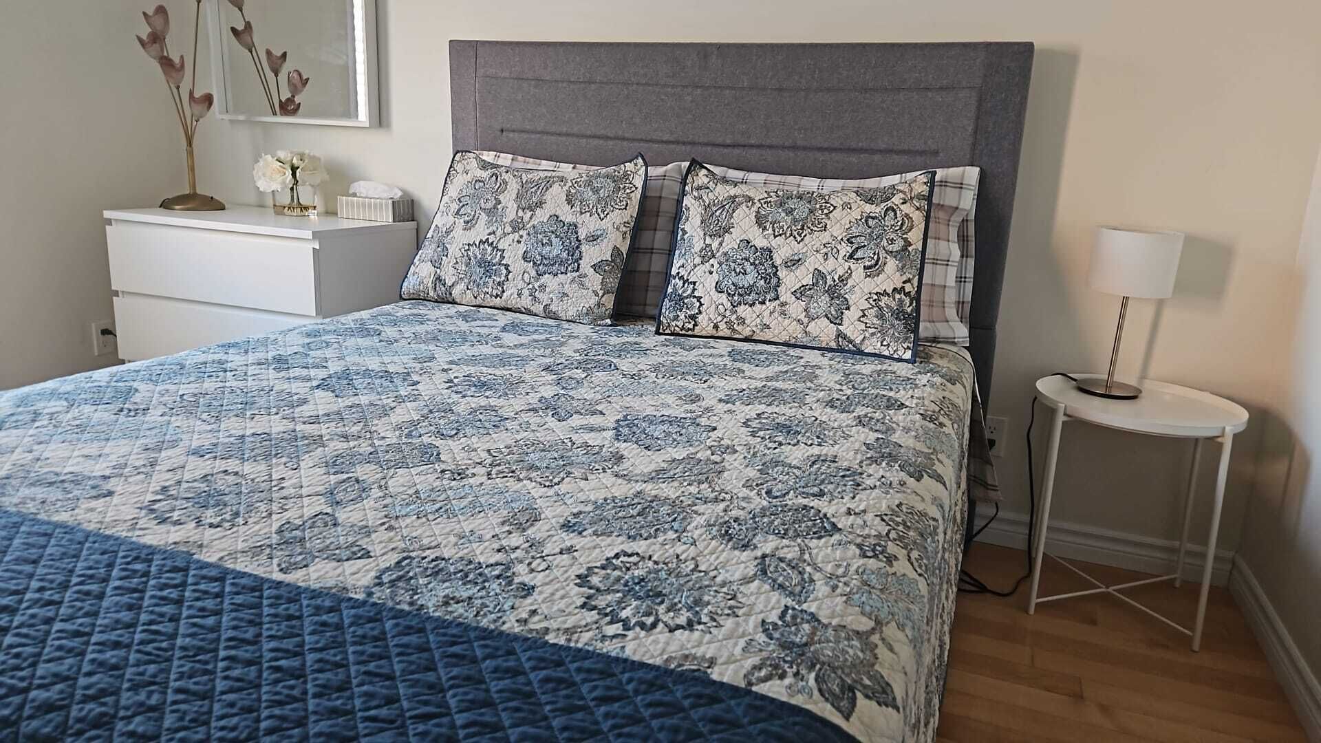 Un lit bien fait, avec une couvre-lit à motifs et deux oreillers, une table de chevet avec une lampe et un miroir fixé au mur.