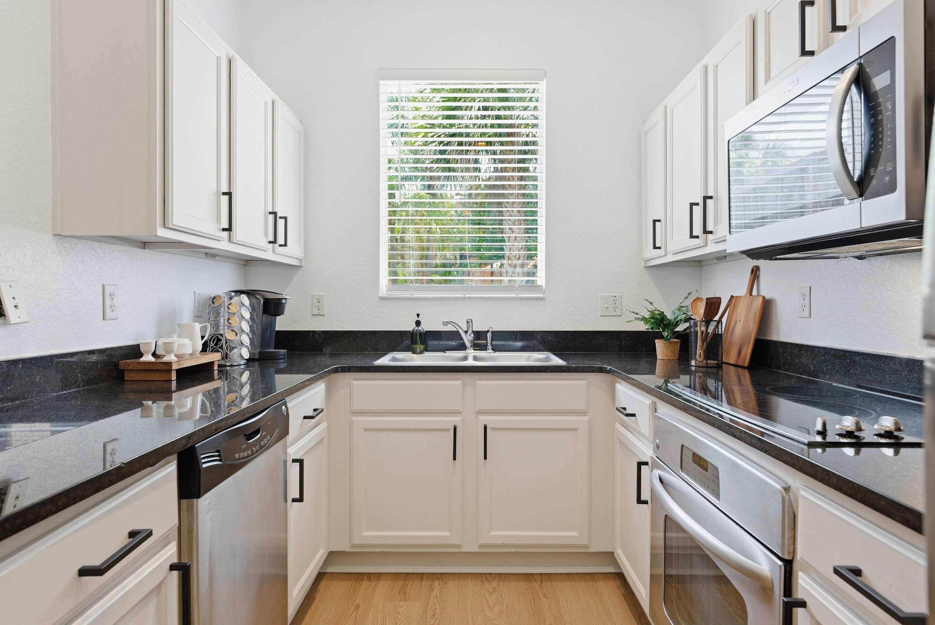 A kitchen with white cabinets, a black countertop, a stainless steel oven, and a window with blinds.
