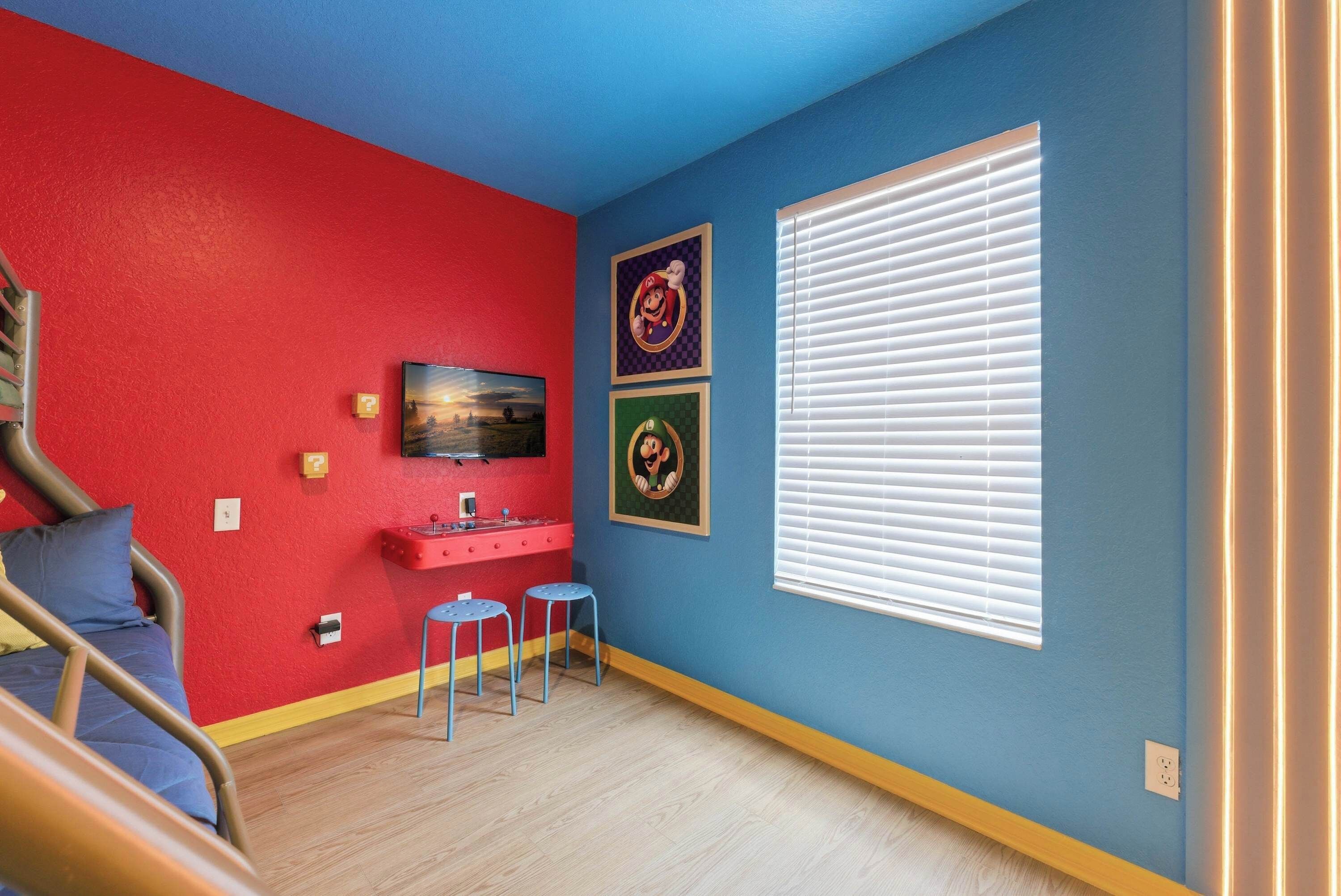 A room with a red wall, blue wall, and a window with blinds. There is a bed, a small table, and two stools.