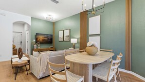 A room with a round wooden dining table, light-colored chairs, a sofa, and a bed. There is a painting on the wall and a lamp on a side table. - Private Jacuzzi, 15 Min to Disney! Modern 4BR Home (Kissimmee)