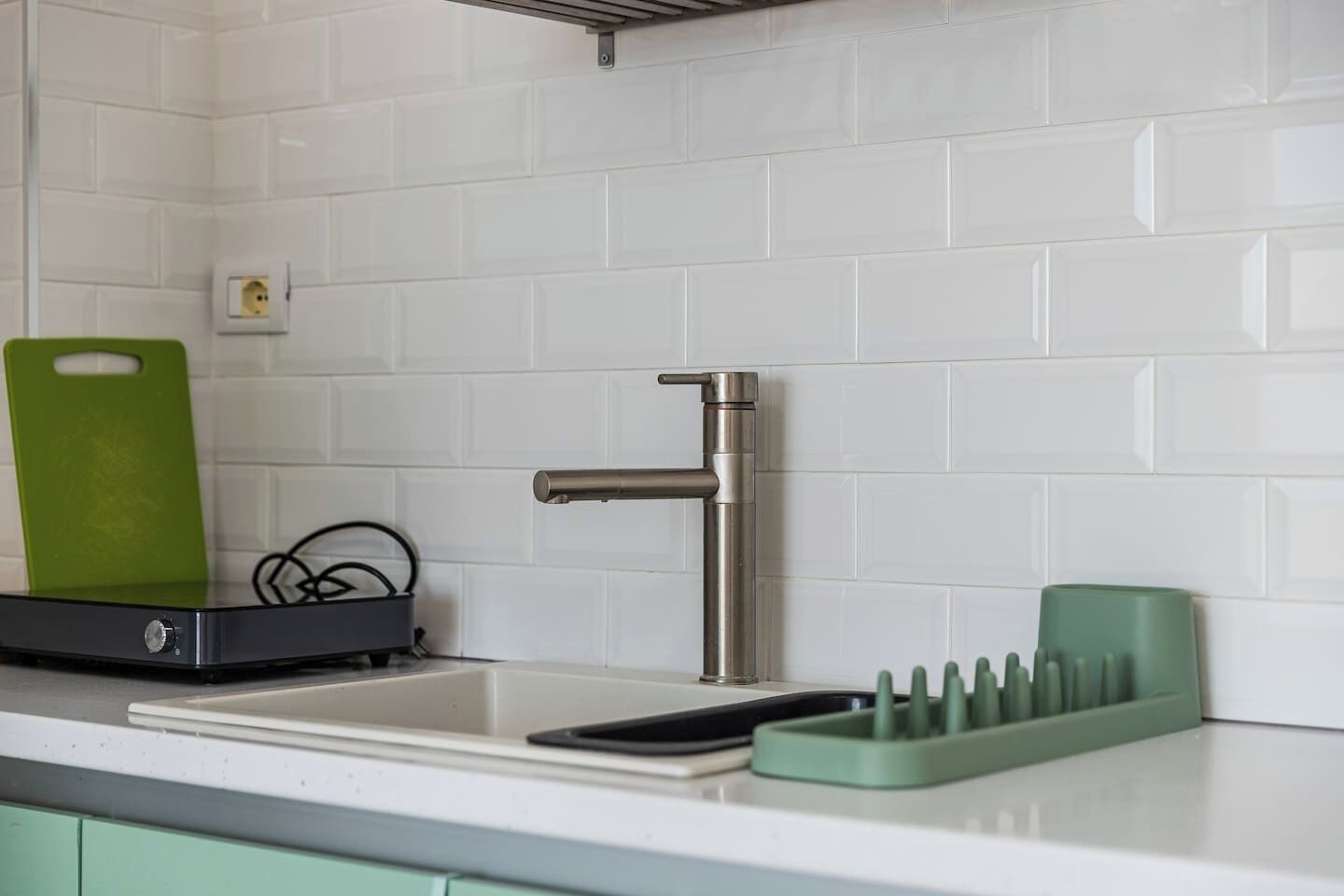 A kitchen with a stainless steel faucet, a green cutting board, a black electronic device, and a green utensil holder.
