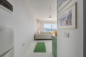 En smal hall med en grön matta, en vit soffa med kuddar, ett fönster med utsikt över havet och inramade tavlor på väggen.