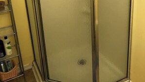 En dusch i ett hörn med duschväggar av frostat glas, en hylla i metall för toalettartiklar och en hörnvägg.