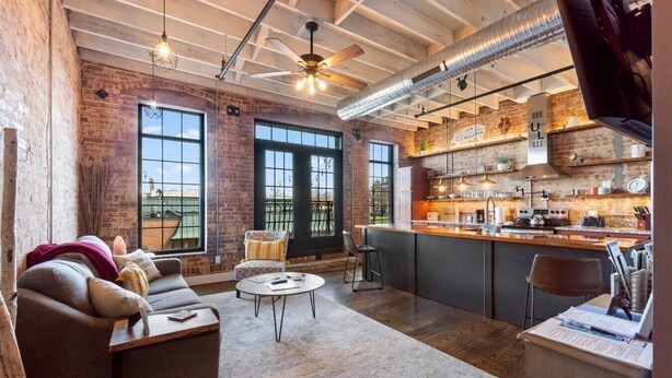 A spacious loft with exposed brick walls, large windows, a kitchen with bar seating, and a ceiling fan. - Downtown Urban Loft above Restaurants (Perryville)