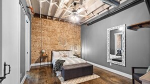 A bedroom with a brick wall, a bed with a headboard, a large mirror, and a ceiling fan. - Downtown Urban Loft above Restaurants (Perryville)