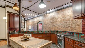 A kitchen with a rustic brick wall, wooden cabinets, a large island, and a built-in oven. - Historic Riverfront Condo for 8, Near Broadway! (Nashville)