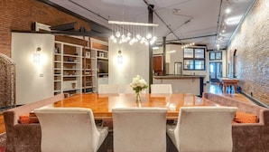 A spacious dining area with a large wooden table, white upholstered chairs, and a hanging light fixture. - Historic Riverfront Condo for 8, Near Broadway! (Nashville)