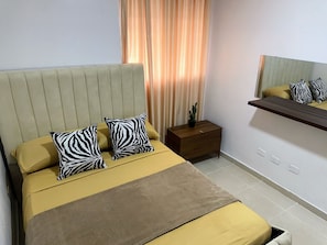A bed with a headboard, two pillows, a bedside table with a plant, and a mirror on the wall.