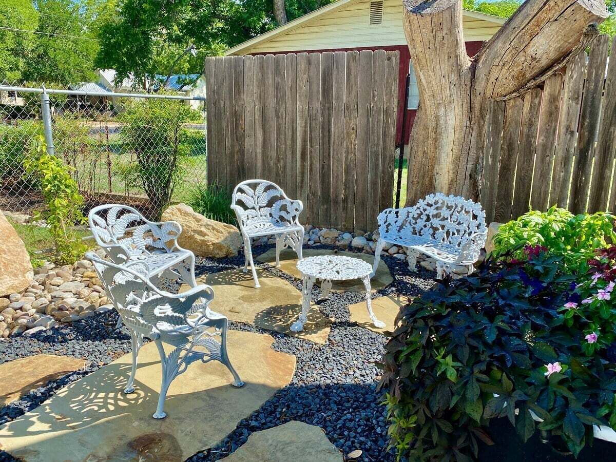 Une terrasse extérieure avec des chaises et une table en métal blanc, entourée d’un chemin de pierre et de verdure.