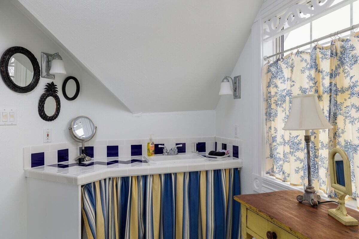 Een badkamer met een wit plafond, een gebogen wastafelmeubel met een spiegel, een wastafel en een raam met patroongordijnen.
