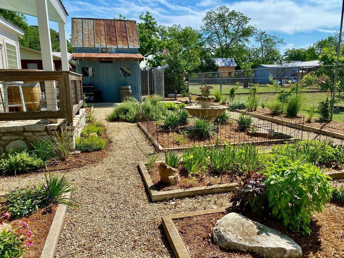 Un jardin avec un chemin de gravier, différentes plantes et un bâtiment de style rustique.