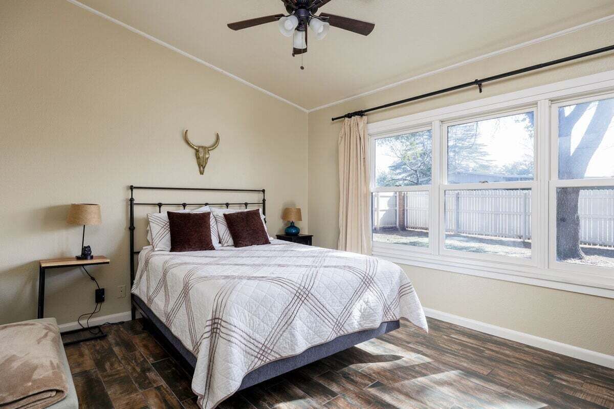 Un dormitorio con cama, ventilador de techo, mesita de noche, lámpara, ventana con vista a una cerca y un adorno de cabeza de toro en la pared.