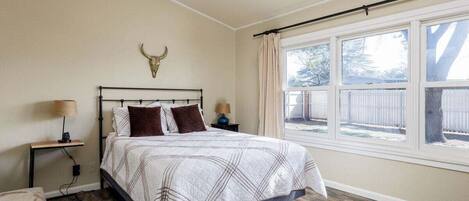 Un dormitorio con cama, ventilador de techo, mesita de noche, lámpara, ventana con vista a una cerca y un adorno de cabeza de toro en la pared.