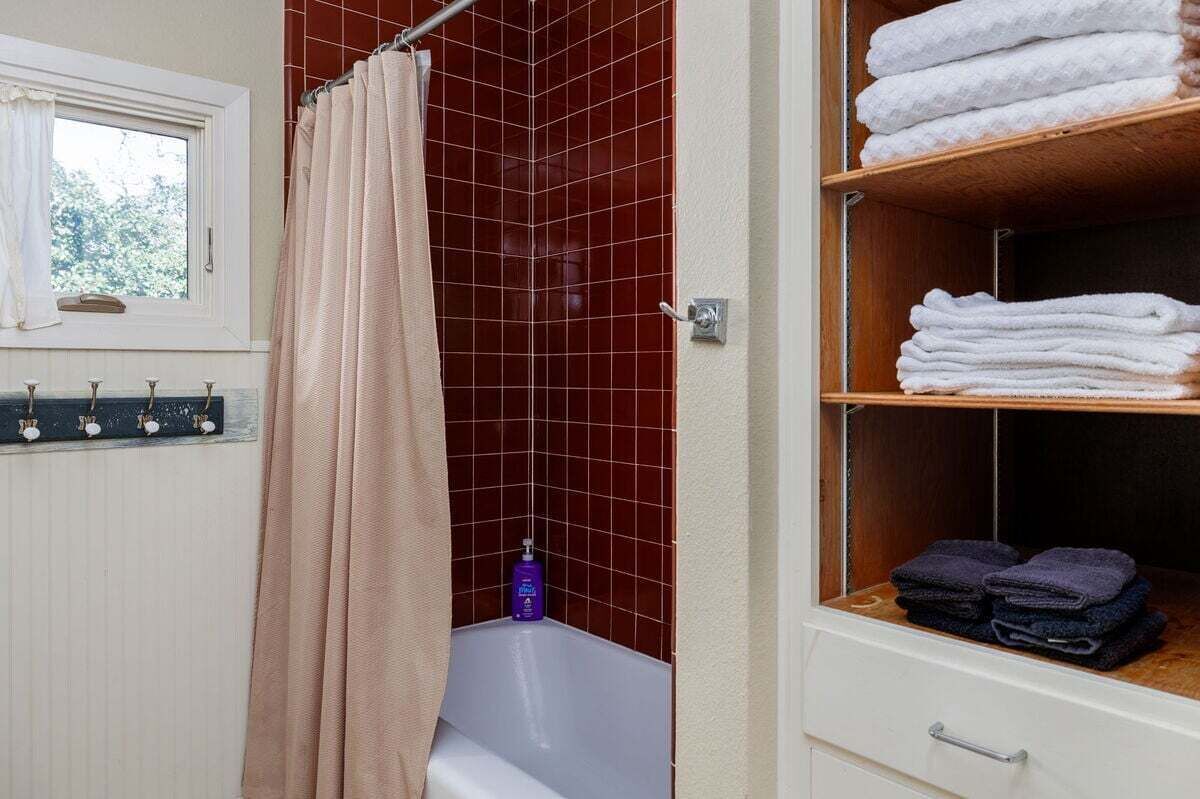 A bathroom with a shower curtain, a bathtub, a shelf with towels, and a window with a view.