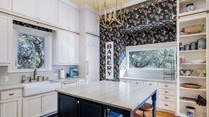 A kitchen with white cabinets, a central island, a 'BAKERY' sign, and a window overlooking greenery. - Unique Victorian Estate Pets Welcome Pool BBQ Picnic Privacy (Dripping Springs)