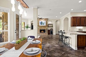 A spacious kitchen with a stone fireplace, wooden cabinets, and a dining area with a long table and bar stools. - Hill Country Escape-670 Heather Hills (Dripping Springs)